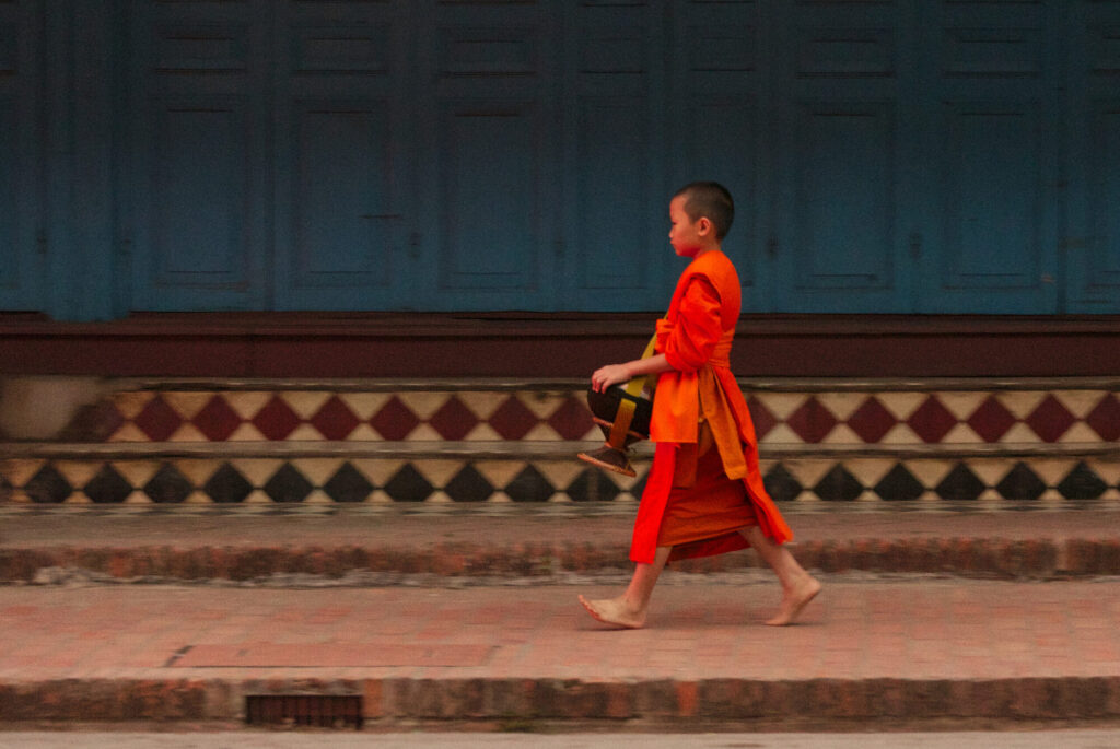 Novice monk walking past a temple wall, almsgiving morning, Laos: Luang Prabang to Vientiane